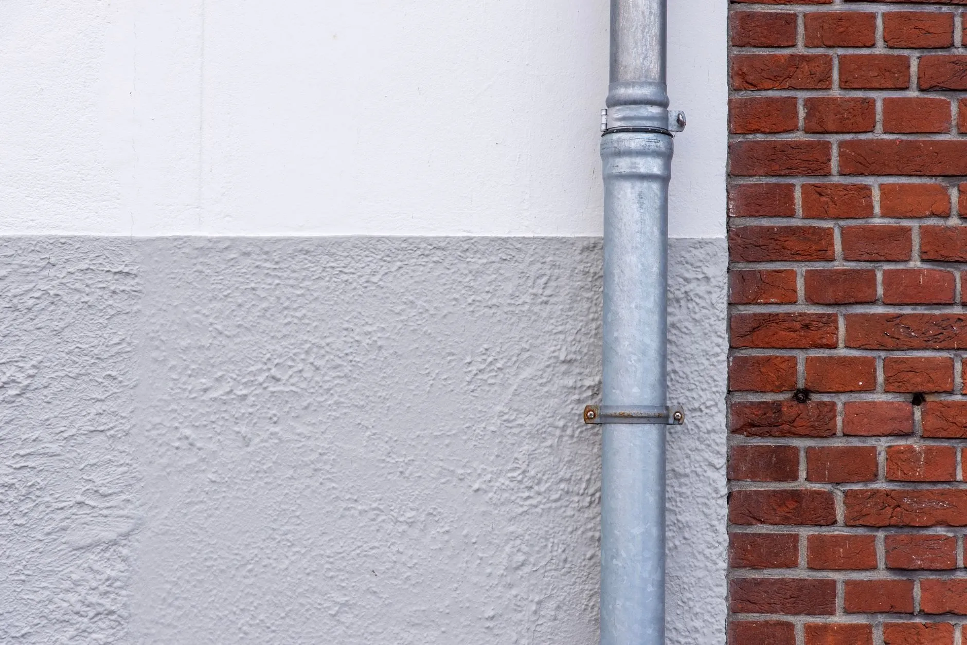 A clean galvanised downpipe fixed to a rendered wall beside red brick