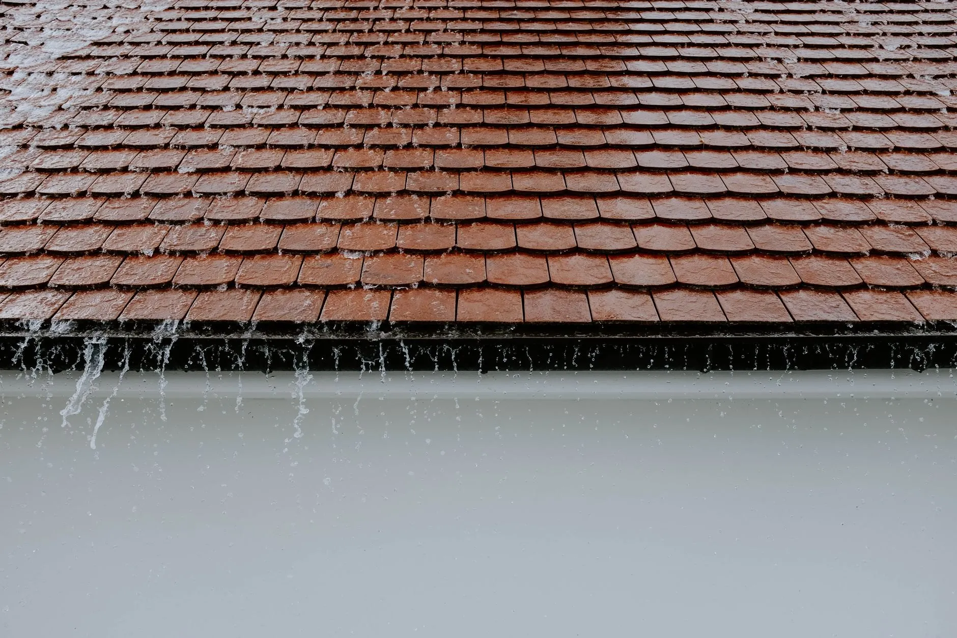 Rain spilling over the edge of a full gutter below a wet clay-tiled roof